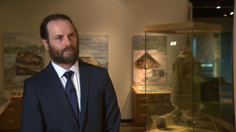 A man with long hair tied into a ponytail, wearing a suit and standing in a museum.