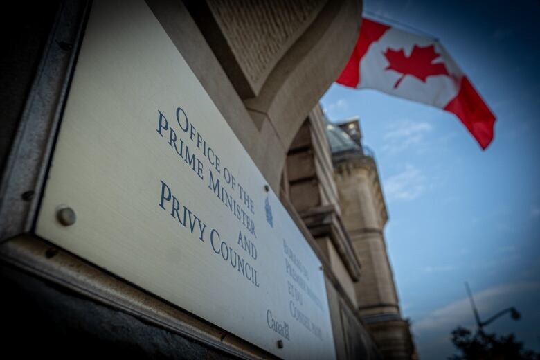 A sign on a stone building says 'Office of the Prime Minister and Privy Council' in English and French. There's a Canada flag behind it.