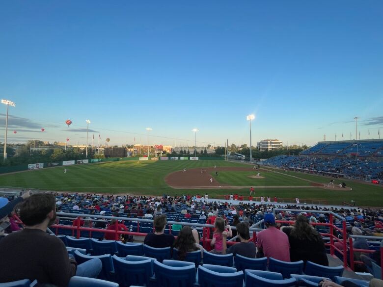 People watch a minor league baseball game in a stadium as the sun sets. There are hot air balloons in the sky in the background.