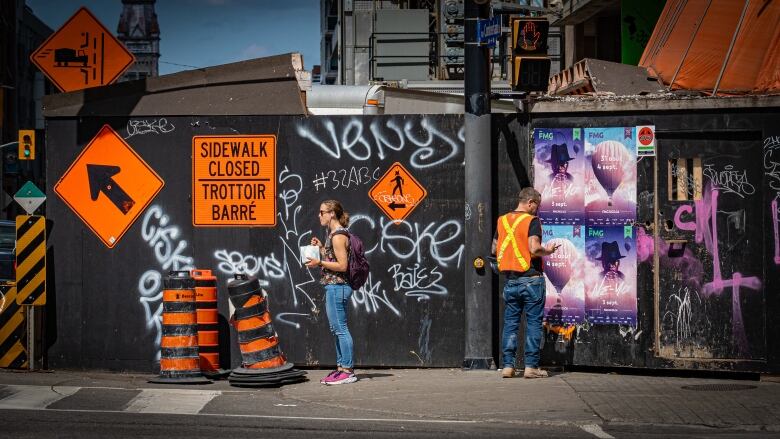 Two people stand by construction fencing on a summer day. The black fence has posters and graffiti on it.