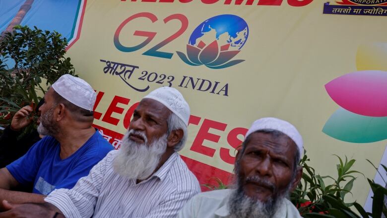 Three elderly men sit in front of a sign welcoming people to the G20 Summit in India.