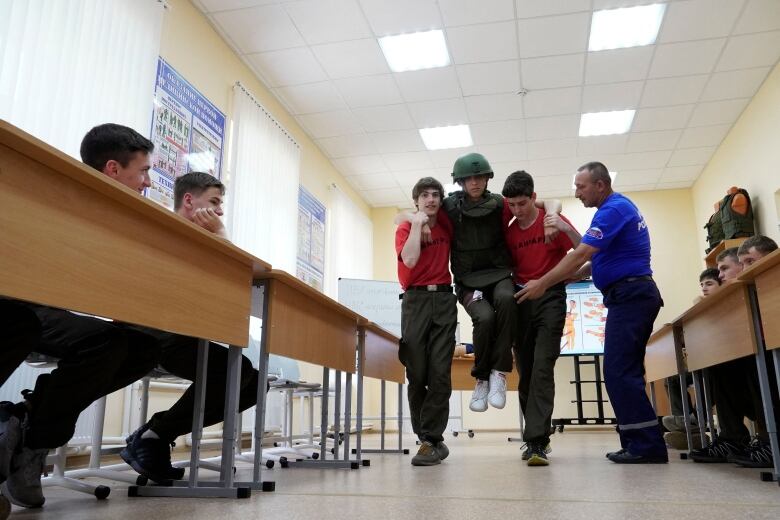 In a classroom, two teenagers carry a third teenager acting like he is injured as other students and a teacher watch.