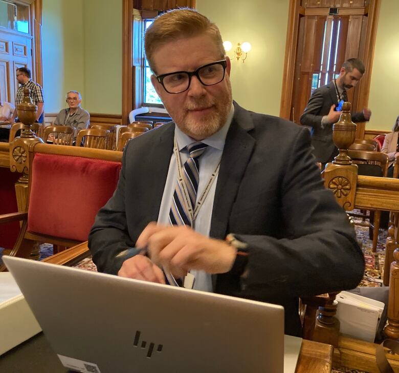 A man with short greying hair opens a water bottle while sitting in front of an open laptop.