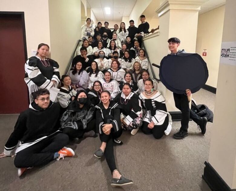 A group of Inuit youth dressed in white and black Inuit hooded parkas sit in a stairway for a class photo.