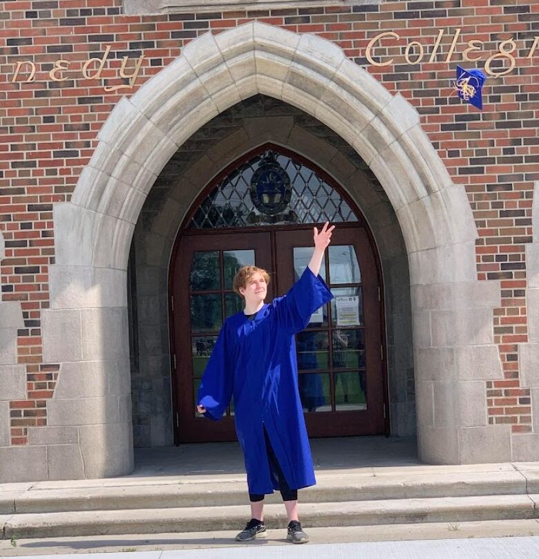 Ashtyn is standing in a blue graduation gown throwing his graduation cap into the air in front of the high school. 