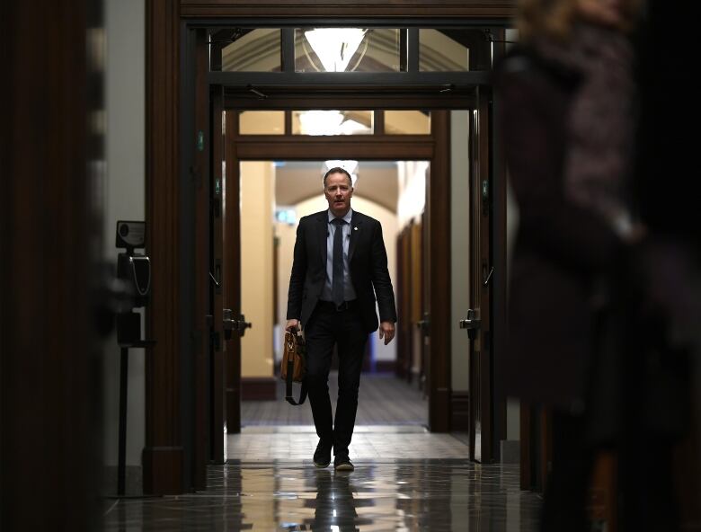 A white man wearing a suit carries a carrier bag through a hallway.