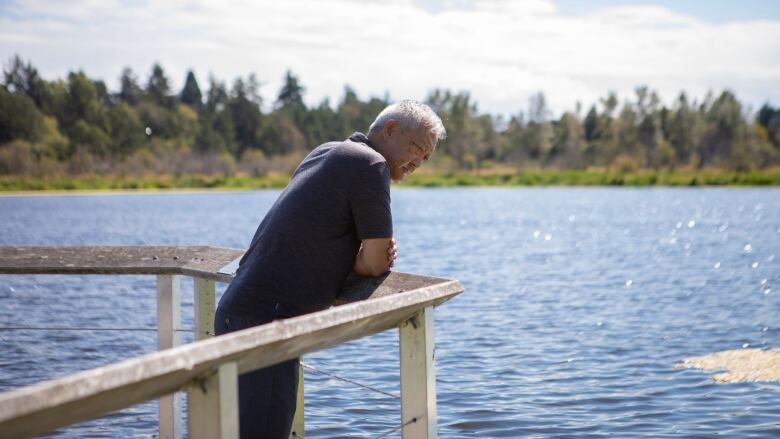 A man leans on a dock railing above open water, a tree-lined shore in the background.