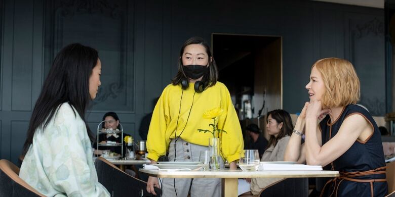 Lulu Wang in a yellow top directs two actresses sitting at a table in a restaurant.