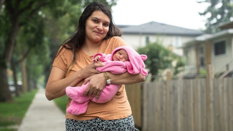 A woman holding a baby stands on the sidewalk.