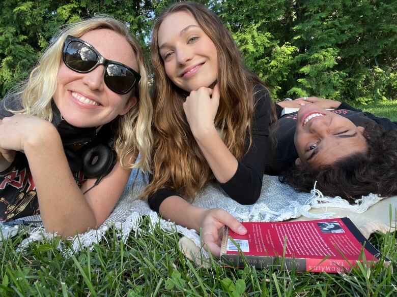 A selfie of Molly McGlynn, Maddie Ziegler, and D'Pharaoh Woon-A-Tai lying on a picnic blanket in the grass, Maddie holding a copy of The Handmaid's Tale.