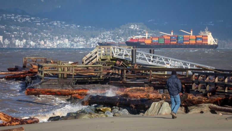 A man walks past a ruined pier on a beach.