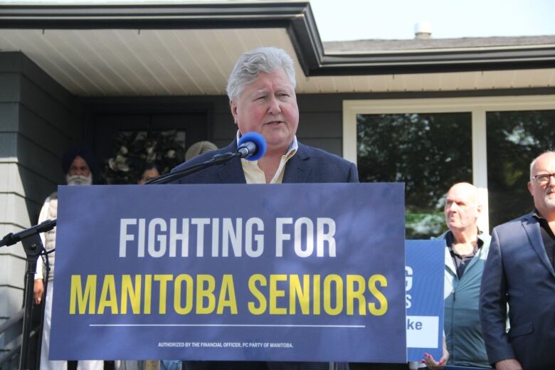 A man in a blue suit speaks at a news conference. The podium has a 'Fighting for Manitoba Seniors' banner on it. 