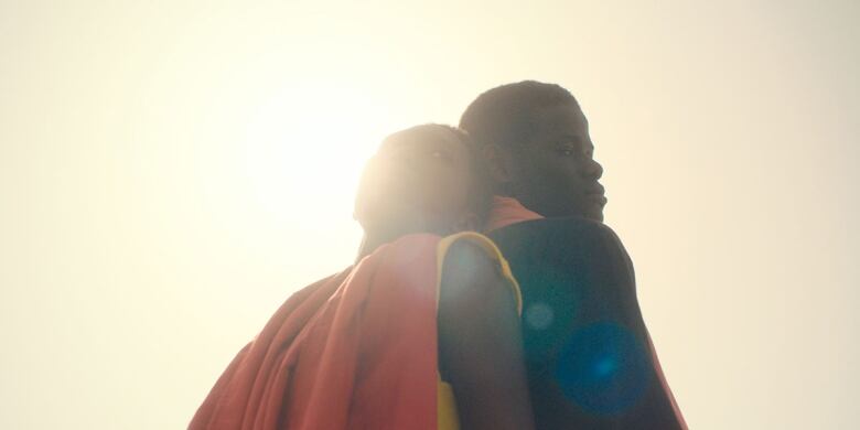 Still frame from the film Banel & Adama. Khady Mane and Mamadou Diallo stand back to back in colourful clothing, gleaming in the sunlight.