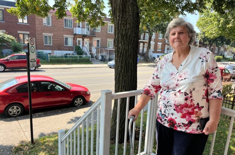 A woman with grey hair, a floral shirt and a cane stands on a balcony in front of a reserved parking spot.