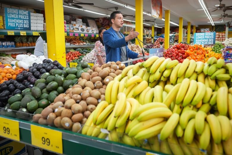 A man in blue blazer gestures at a lectern, surrounded by a grocery produce section.