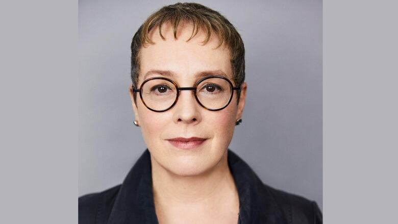 Woman with short hair and bangs wearing tortoise framed glasses and a navy blue collared shirt stands in front of grey background.