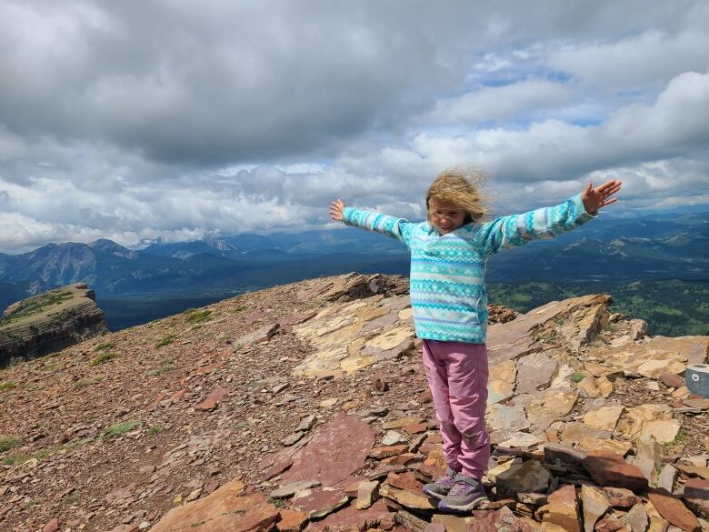 A child is seen standing among rocks at the top of a mountain with peaks and cloudy blue skies in the background.