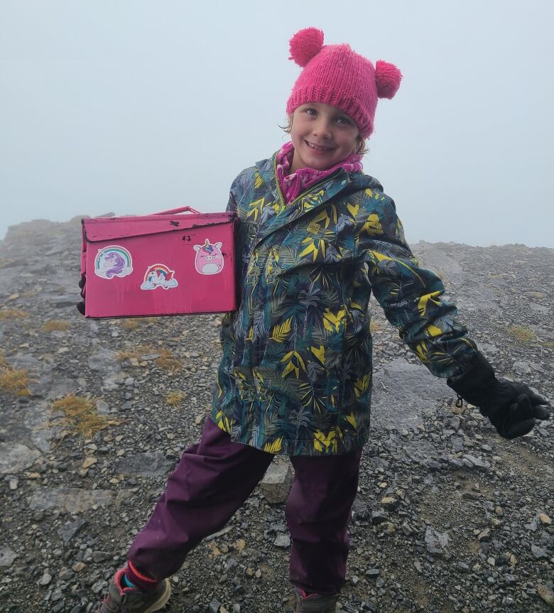 A child in a dark green rain jacket and purple pants is seen posing with a pink summit register.