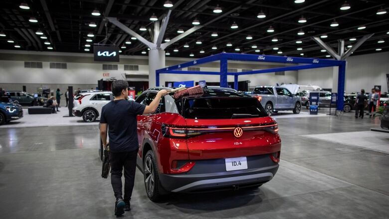 A sleek red vehicle is on display under a ceiling full of recessed pot lights in a cavernous indoor space. Other cars are visible in the background.