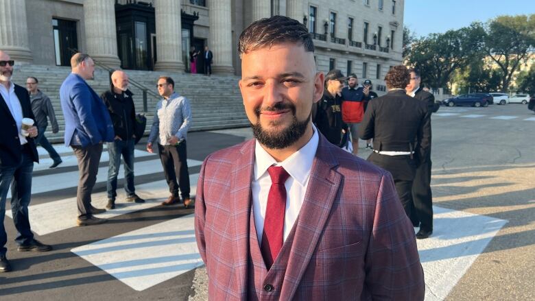 A main wearing a suit stands outside a building. Some people stand behind him.