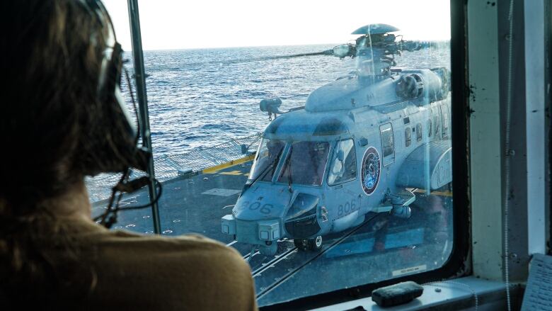 A sihouetted person wearing headphones looks through a window at a helicopter that is landing on a ship deck. 