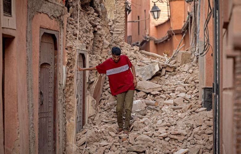 A man in a red shirt leans against a wall as he walks in a narrow alleyway filled up with rocks.