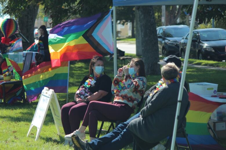 Several people wearing masks sit together on folding chairs outside. Pride flags can be seen around them.