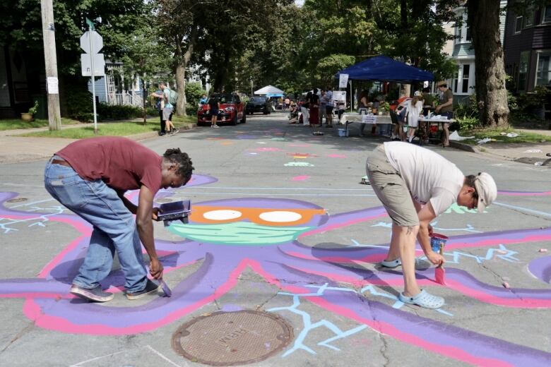 Two people are seen paiting a large purple octopus on a city street.