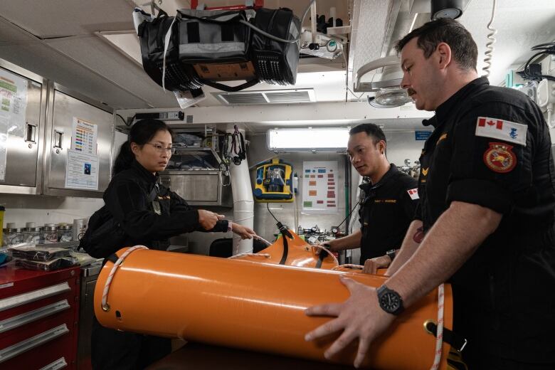 A woman and two men, all in navy uniforms, unroll an orange synthetic sheet of material while inside a cramped medical office. 