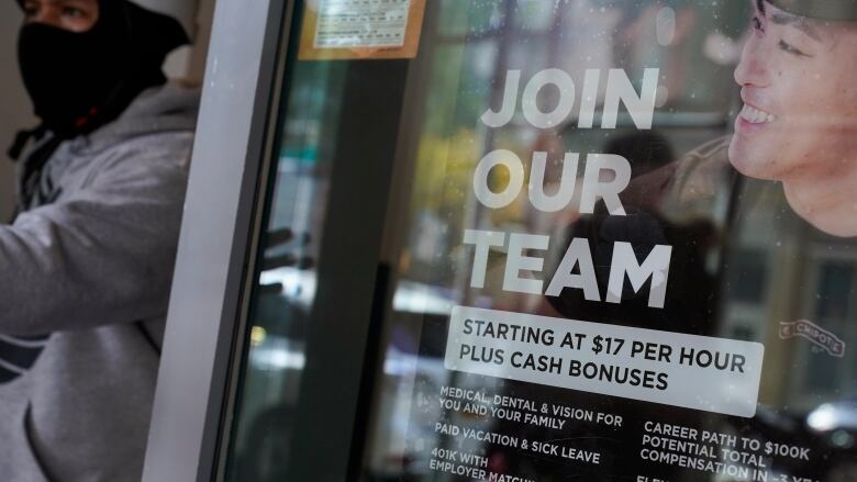 A job posting sign hangs in the window of a fast-food restaurant, as a man wearing a mask and a helmet exits. It reads: 'Join our team. Starting at $17 per hour plus cash bonuses.'