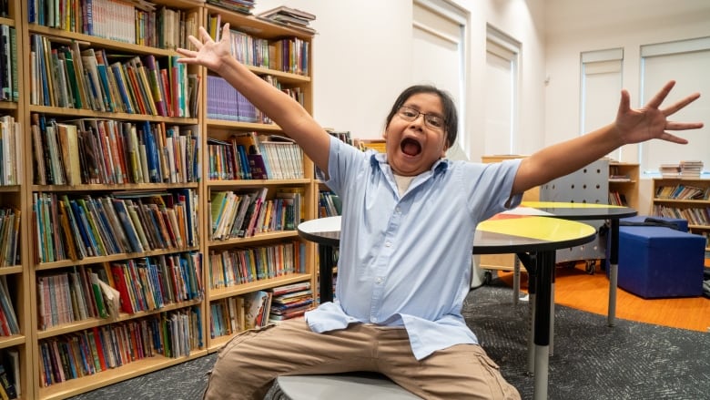 A young person sits on a chair and waves their arms in the air. Behind them are bookcases.