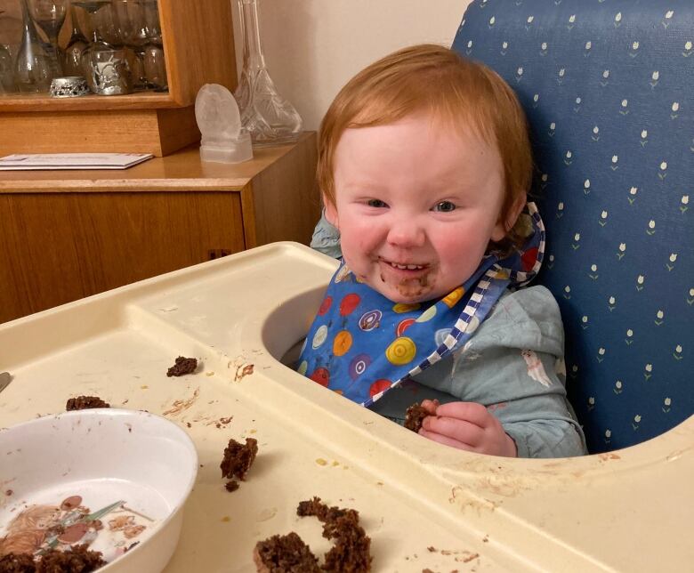 A baby in a highchair looks at the camera. She has meat sauce all over her face, as well as on the highchair table.