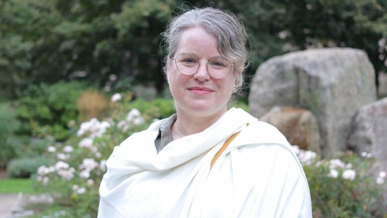 A person with mid-length hair and glasses looks at the camera with a neutral expression. Behind them is a garden with flowers and rocks.