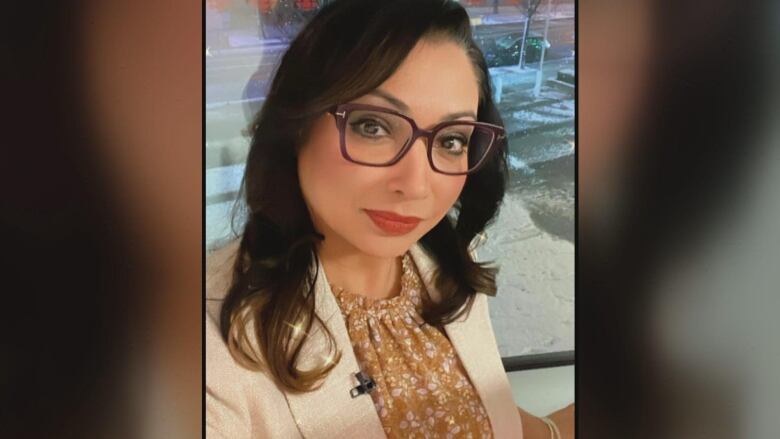 A South-Asian woman of about 40 years old poses for the camera in a beige blazer and mustard-coloured blouse. She has shoulder-length brown hair and wears black-rimmed glasses.