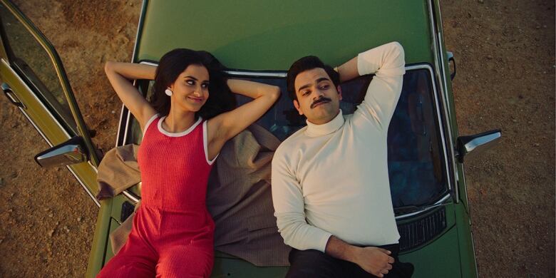 Still frame from the film The Queen of My Dreams. Amrit Kaur smiles at Hamza Haq as they lie on top of a green car.