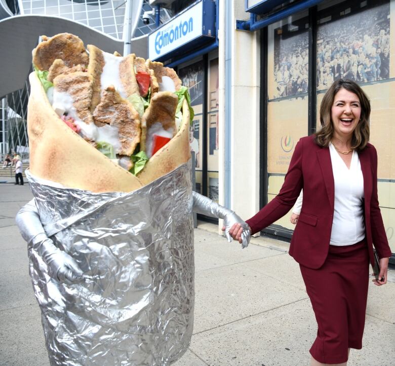 A life-sized donair with arms walks hand in hand with a woman in maroon power suit.