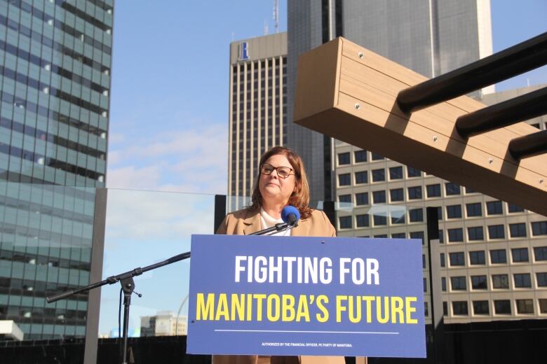 A woman speaks behind a podium, on the second-floor of an outdoor terrace as downtown skyscrapers are seen behind. 