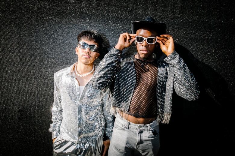 An East Asian man and a Black man, clad head-to-toe in silver, look at the camera.