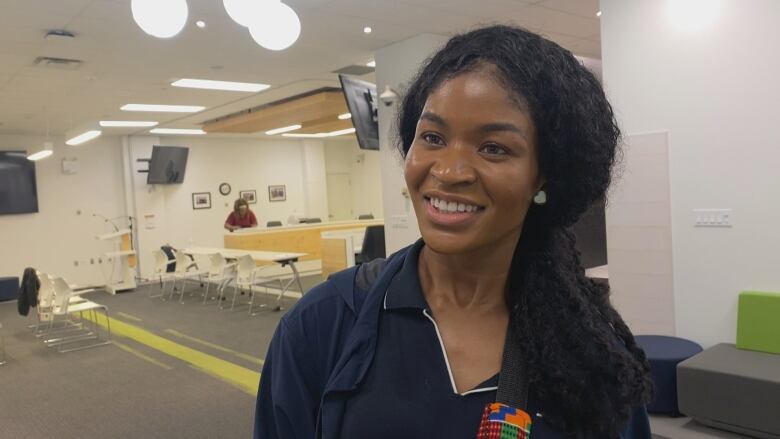 A young Black woman with her long black hair in a ponytail falling to the side wears a navy shirt and jacket as she stands in a large open room with white walls