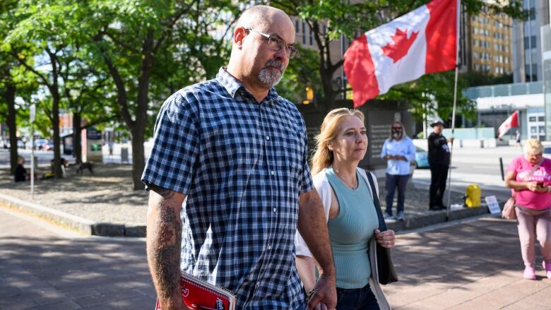 A man holds hands with a woman as they walk to a courthouse. Someone holds a Canada flag in the background.