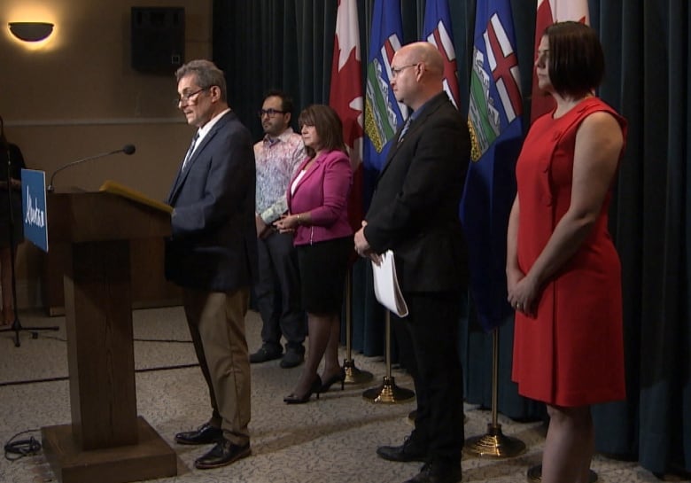 A man address the media from a podium, while four other people look on.