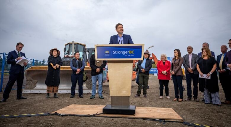 A man speaks from a podium that says StrongerBC flanked by more than 10 dignitaries, including MLAs. At the far left stands Health Minister Adrian Dix.