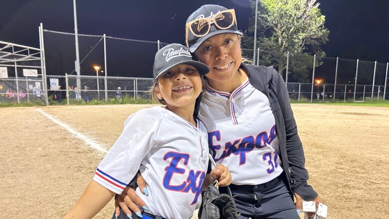 A young girl and her mother, both dressed in baseball uniforms, pose for a photo.