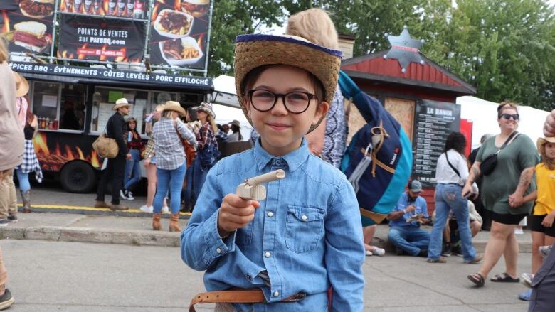 A little kid standing on a busy street wears a blue button-up shirt and points a wooden pistol at the camera. He is wearing glasses and a coyboy hat. 