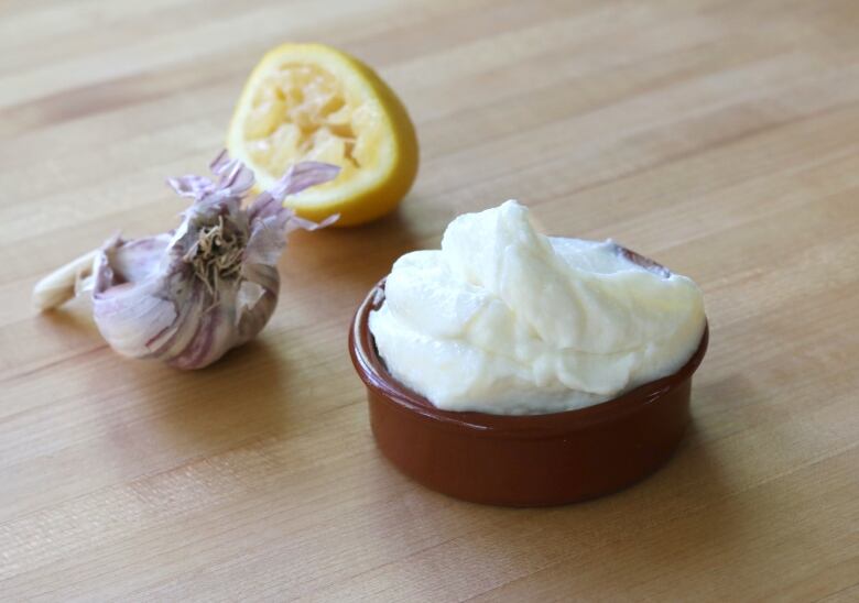 A container filed with creamy Lebanese garlic sauce called toum next to a garlic bulb and lemon.