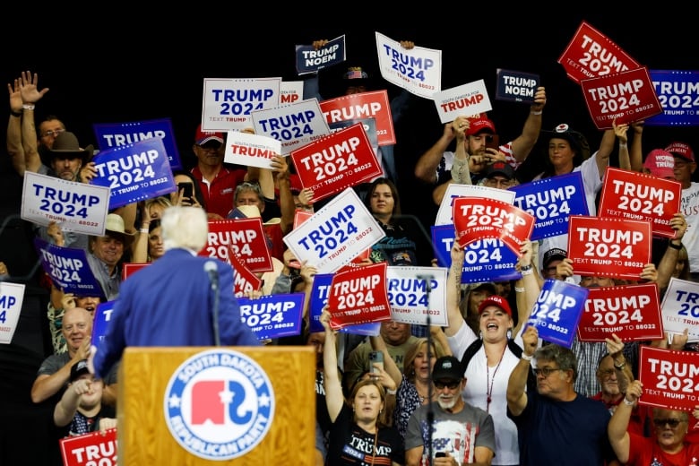 Trump, seen from behind, speaking to crowd of supporters holding posters positioned behind a stage