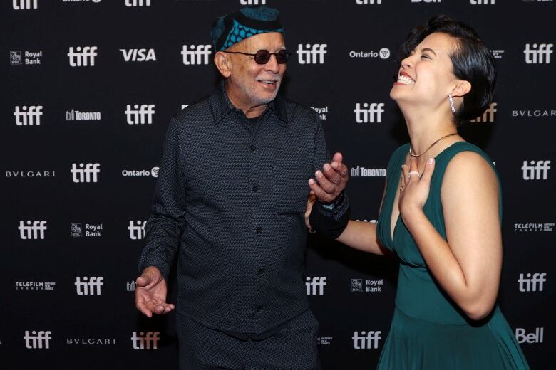 A woman in a green dress with plunging neckline holds one hand to her chest and tosses her head back laughing, while her other hand holds the arm of a director. Both are walking the red carpet at TIFF.