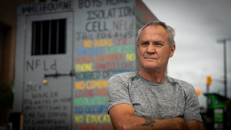 A man wearing a grey t-shirt with his arms folded. He's standing in front of a grey box with multicoloured lettering on it. 