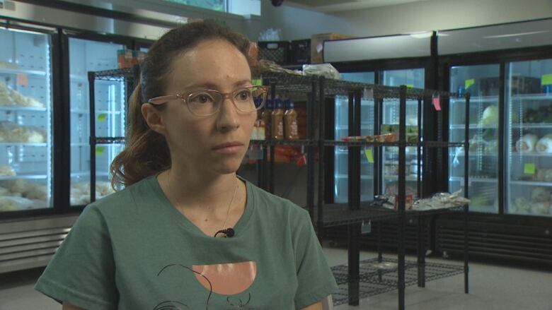 A woman wears glasses and a blueish shirt with a pink pattern on it. She wears glasses and a ponytail. She is sitting in front of black shelves and industrial fridges. 