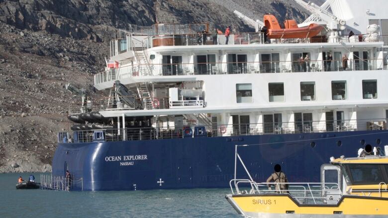 Small cruise ship with steep embankment visible behind and a man looking on from a small boat. 